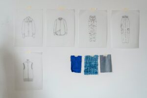 A collection of fashion design sketches with fabric swatches pinned to a white wall in a design studio.